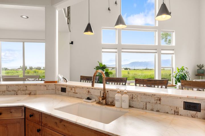 Kitchen sink and counter with pendant lights, looking out tall windows toward fields and mountains