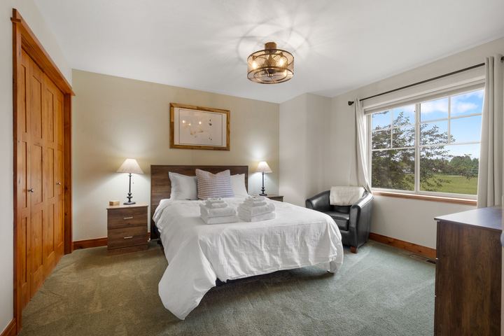 Upstairs bedroom with white bedding, bedside lamps, a leather armchair, and a window over the yard